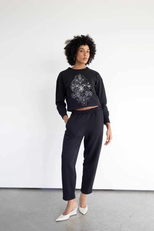 A woman wearing the Baxter Fleece Pants by Eve Lavoie, ankle-length joggers with an elastic waist and side pockets. She is wearing them with the Hanay Sweatshirt in Black and standing in front of a white background. 