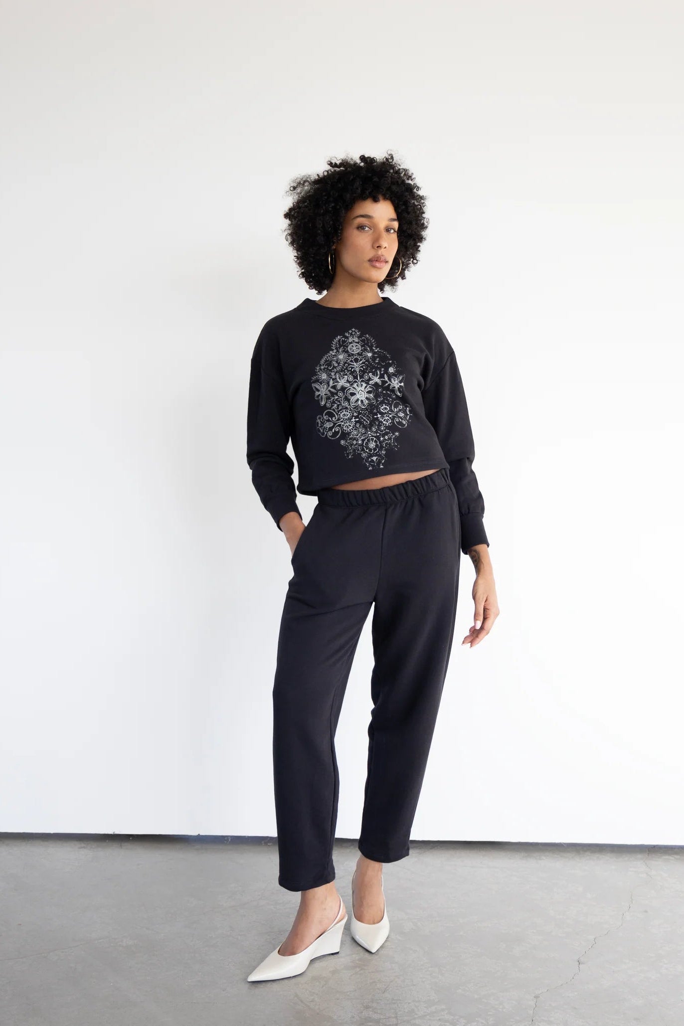 A woman wearing the Hanay Sweatshirt by Eve Lavoie in Black, a slightly cropped sweatshirt with a hand-printed silkscreen design. she is wearing it with black pants and standing in front of a white background. 