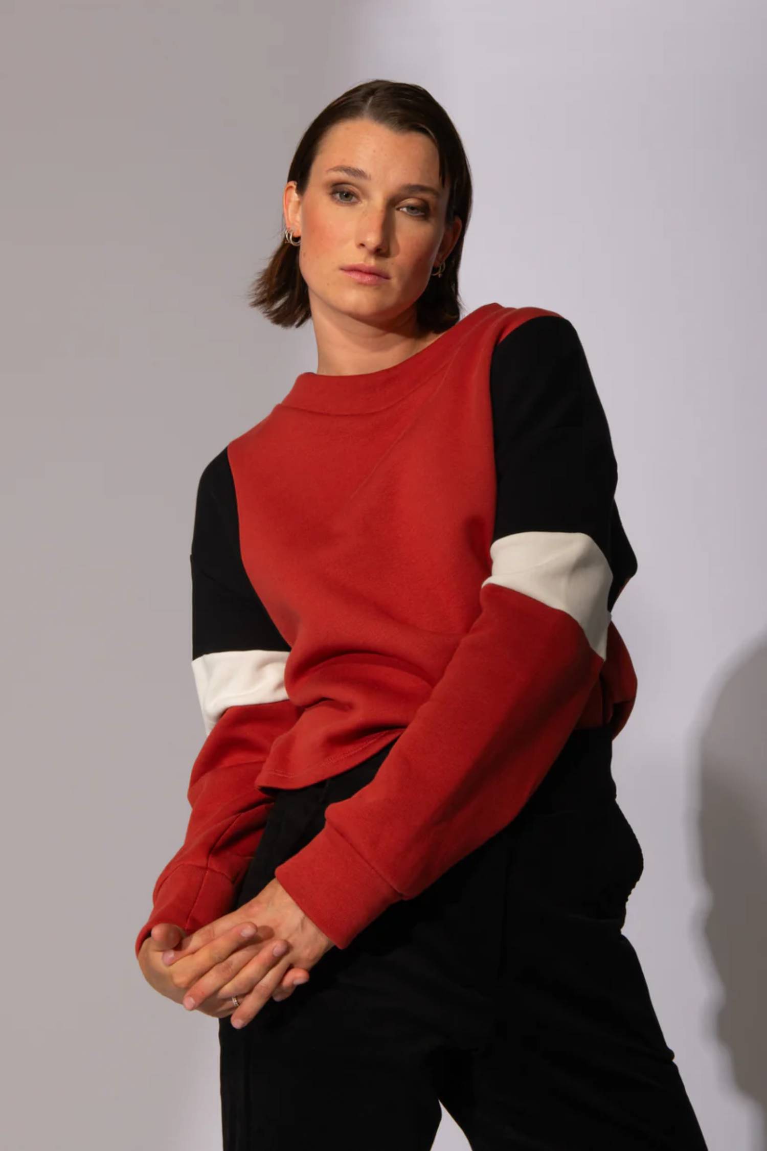 A woman wearing the Eve Lavoie Sour Time crewneck in Terracotta with Tricolour Cutout, featuring black/terracotta/white colourblocking. She is wearing black pants and standing in front of a grey background 