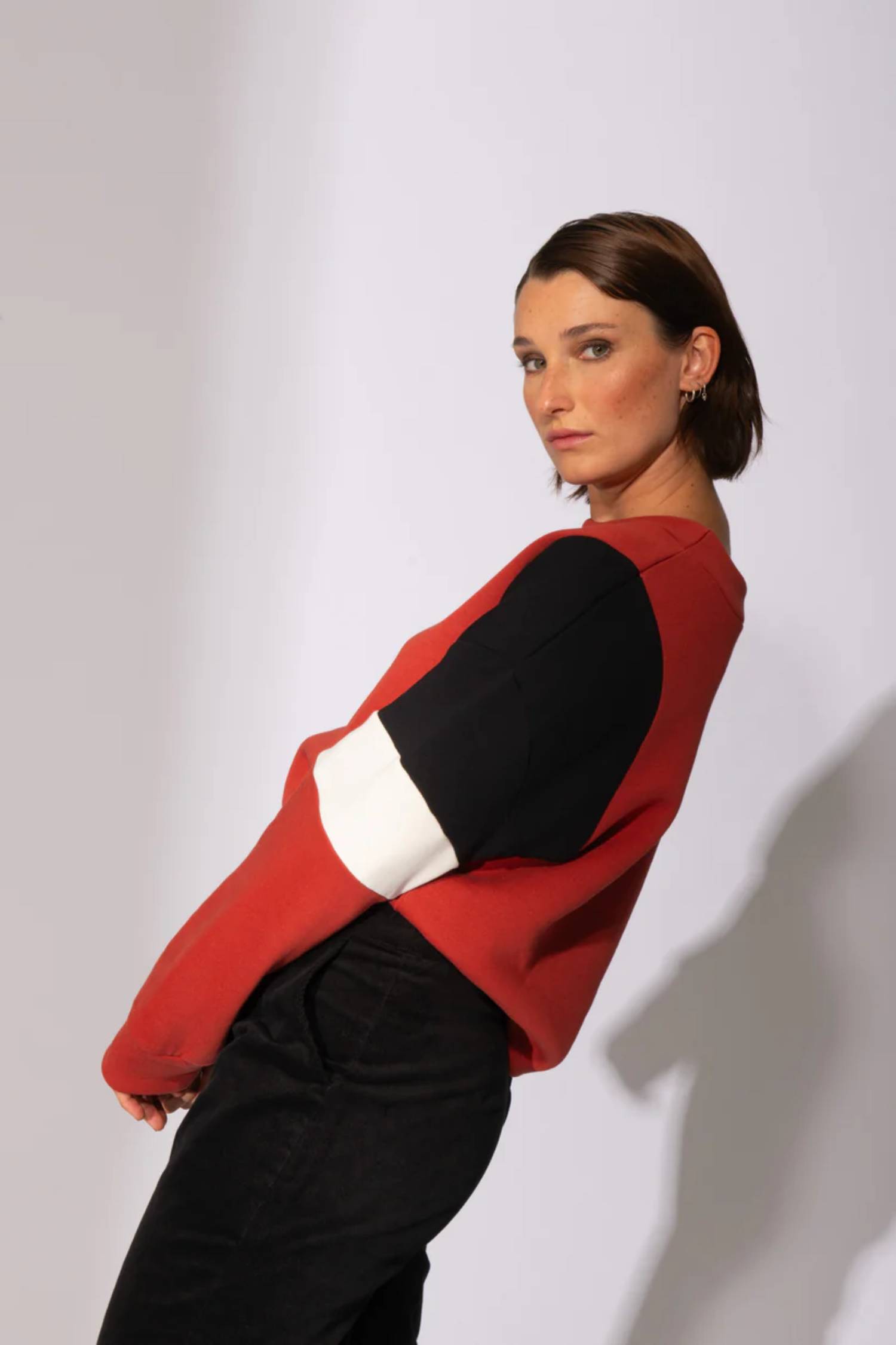 Side view of woman wearing the Eve Lavoie Sour Time crewneck in Terracotta with Tricolour Cutout, featuring black/terracotta/white colourblocking. She is wearing black pants and standing in front of a grey background 