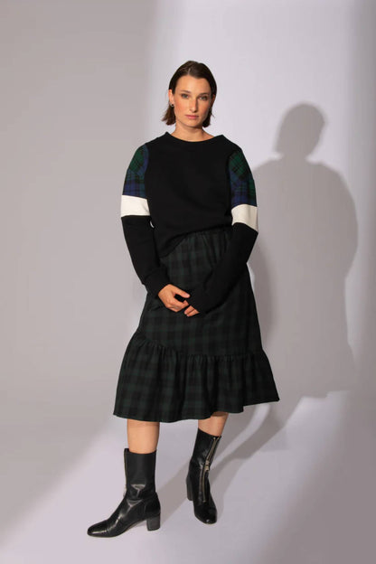 A woman wearing the Eve Lavoie Sour Time crewneck in Black with Tricolour Cutout, featuring tartan/black/white colourblocking. She is wearing a tartan skirt and standing in front of a grey background  