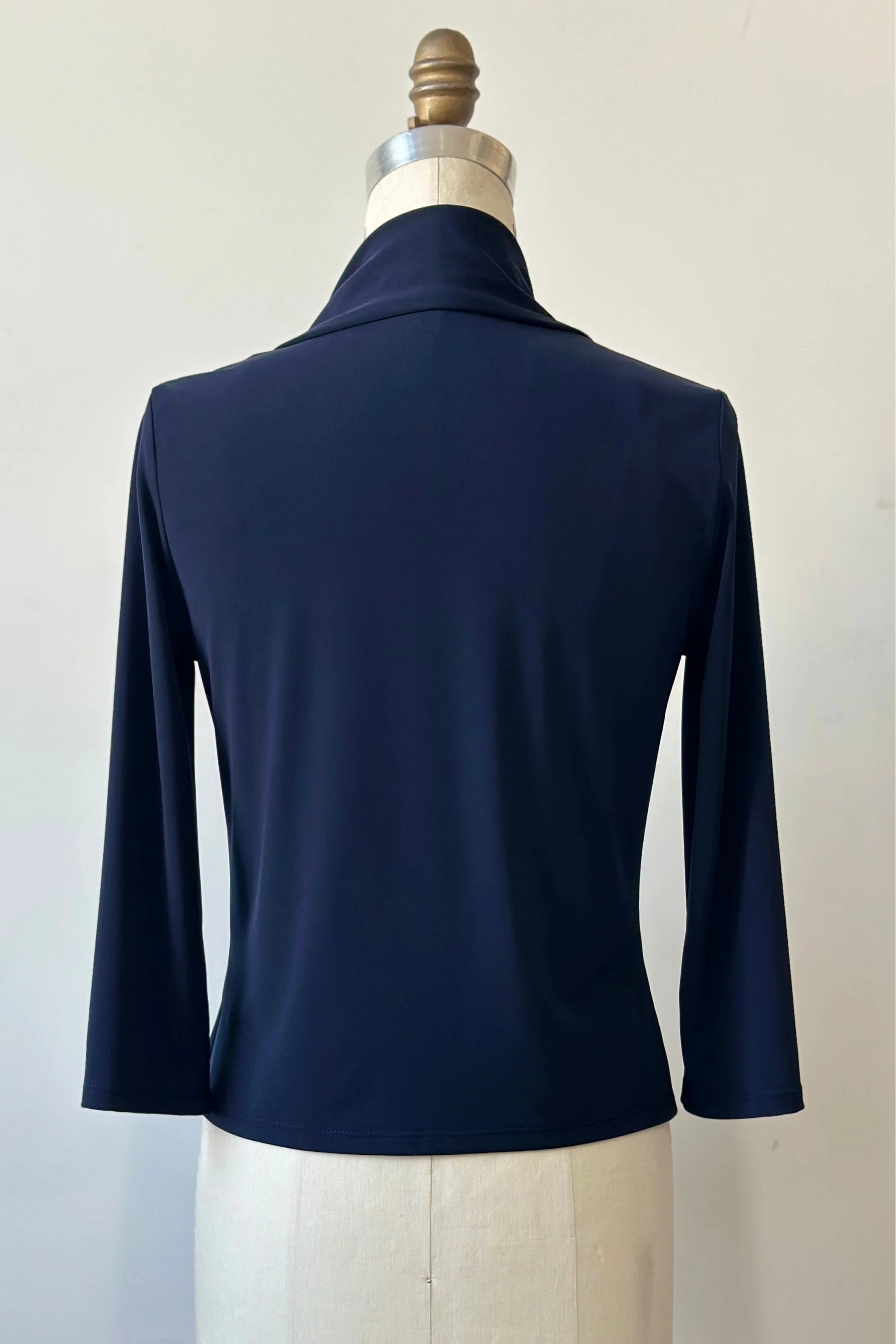 A back view of the Lioren Cardigan by Kollontai in Navy, a stretchy cropped jacket with longer pointed panels in the front that can be worn loose or knotted, is shown on a mannequin in front of a white background. 
