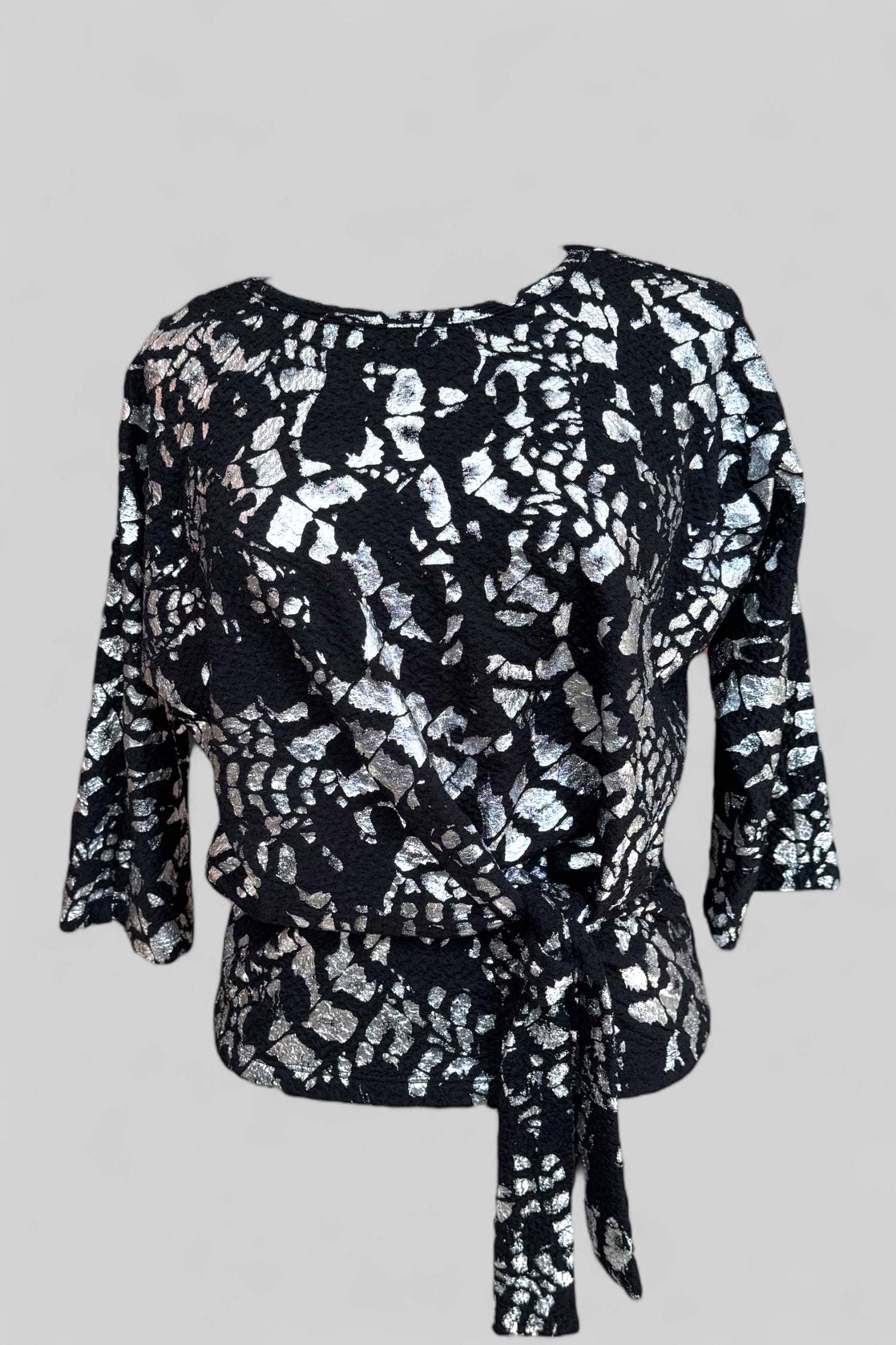 The Posey Sweater from Compli K in Black and Silver, with a round neck, 3/4 sleeves, and a tie detail at the waist, with black pants, is shown against a grey background.