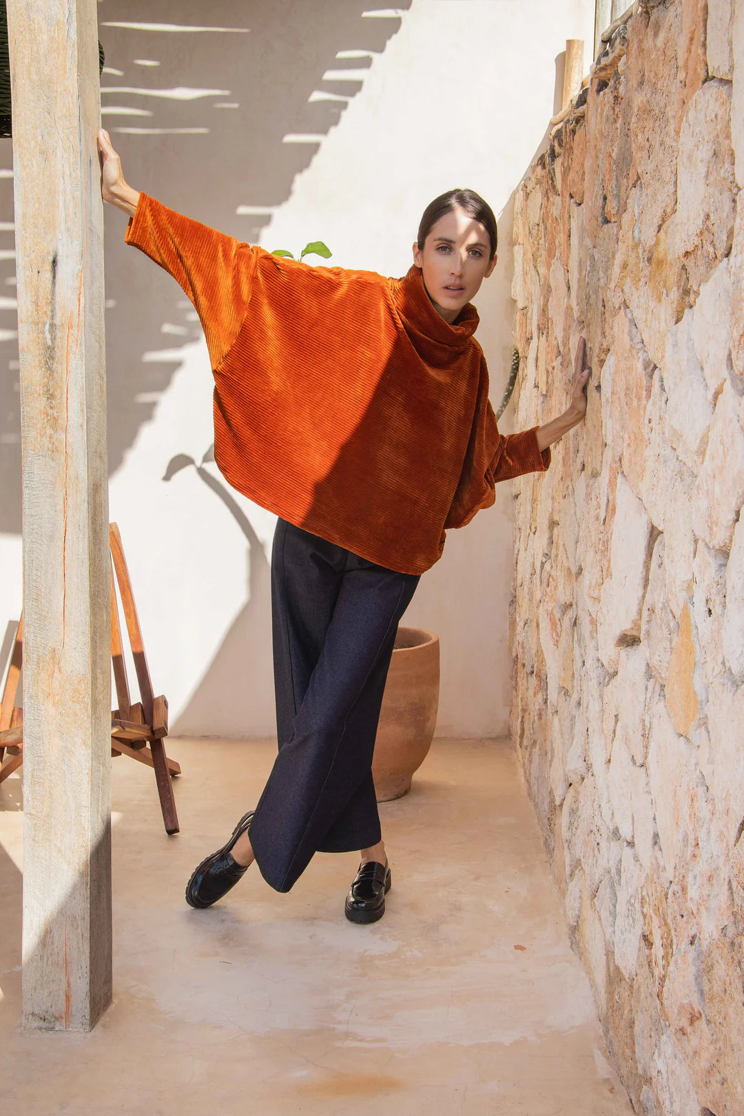 A woman wearing the Vienna Pants by MAS in Jeans, with the Amsterdam Sweater by MAS in Amber, leaning against a stone wall. 