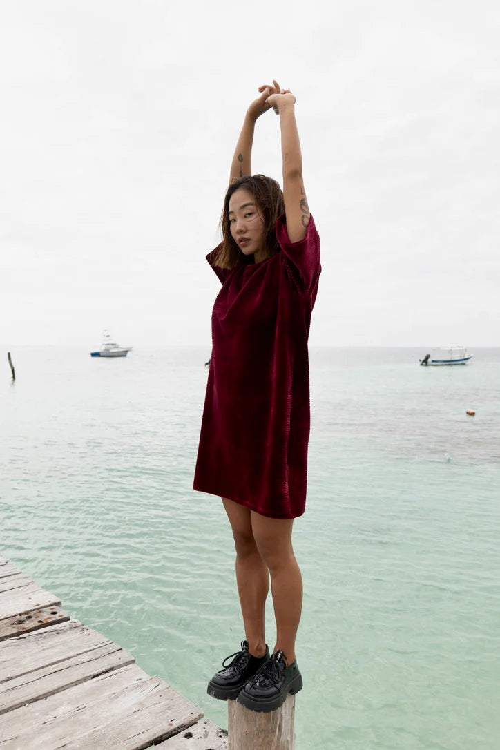 A woman wearing the Mykonos Dress by MAS in Rosewood, a short sleeved boxy, knee-length dress in ribbed velvet fabric. She is standing on wooden dock. 