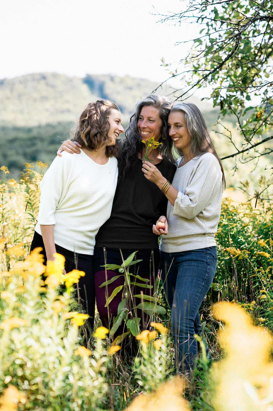 Three women wearing the Perla Top by Marie C,  one in Ivory and one in Black, with a wide round neck, 3/4 batwing sleeves, and fitted waist in ribbed knit fabric. They are standing outside. 