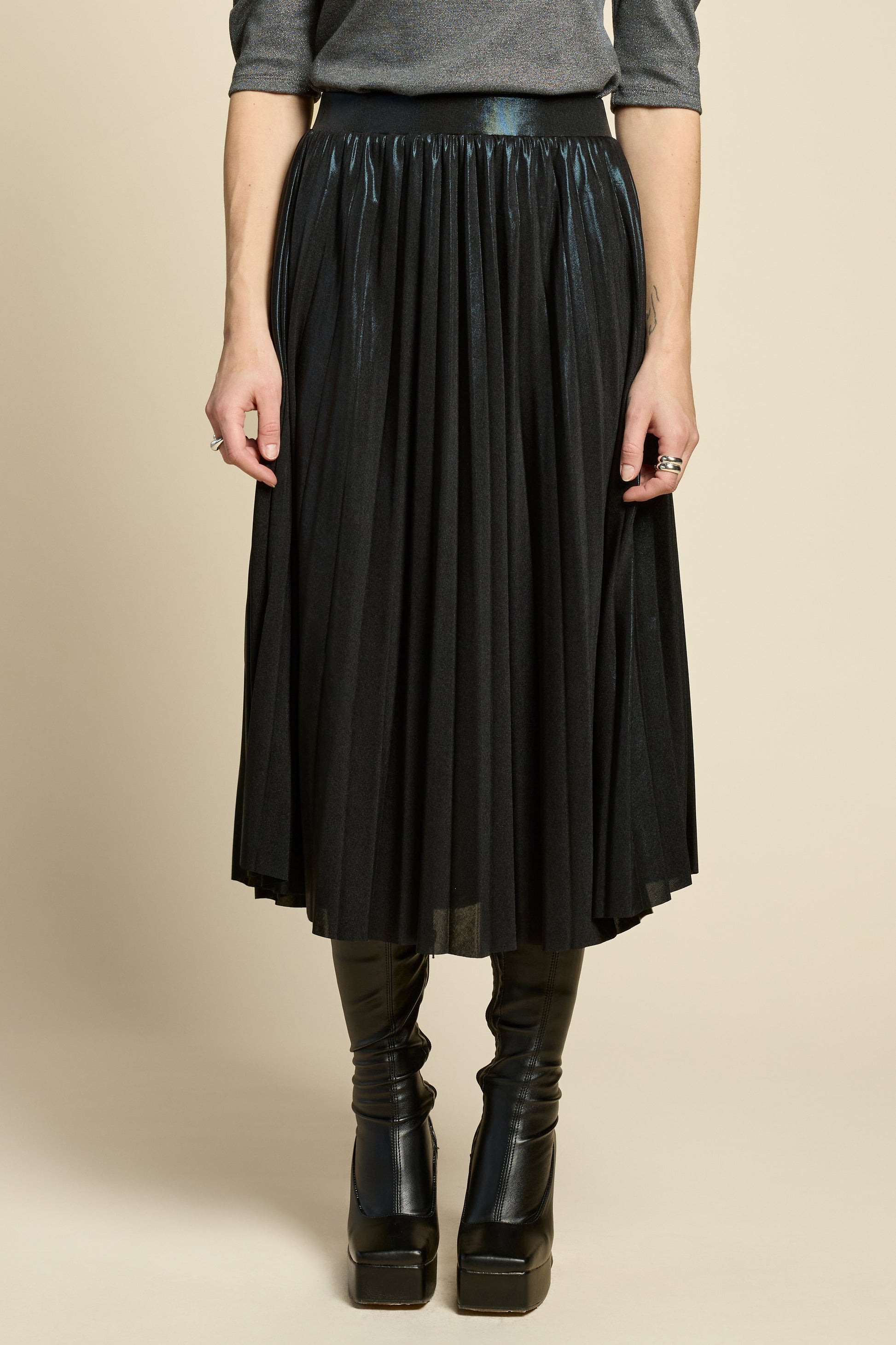 Waist down view of a woman wearing the Feyer Skirt by Cokluch in Glow Knit Black, a midi length pleated skirt in shimmery metallic fabric. She is wearing it with the Insomie top in Facet Black and is standing in front of a beige background. 