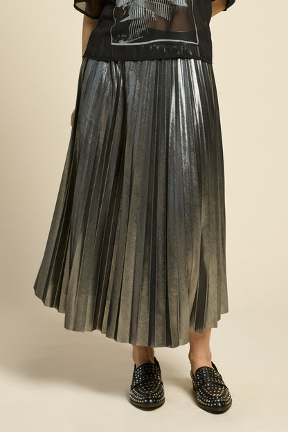 Waist down view of  woman wearing the Feyer Skirt by Cokluch in Glow Knit Silver, a midi length pleated skirt in shimmery metallic fabric. She is wearing it with a sheer black graphic top and standing in front of a beige background. 