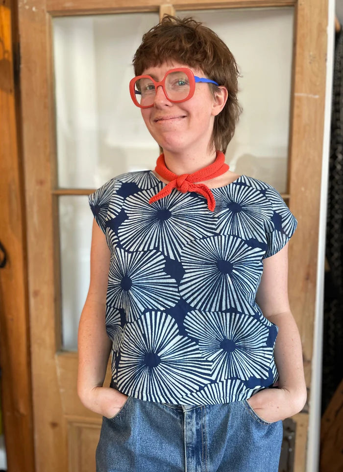 A woman wearing the wearing the Sandrigo Top from Kazak in Indigo Floral, a loose cotton top with short extended sleeves and a wide round neck. She is wearing it light jeans and an orange scarf and standing in front of a wooden door. 