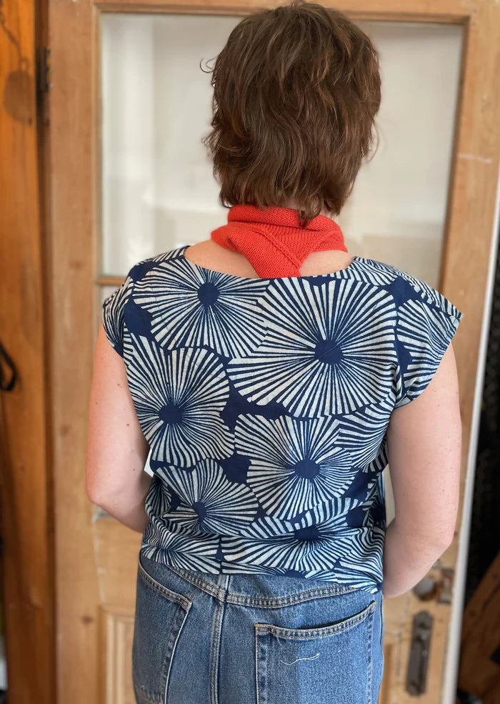 Back view of a woman wearing the wearing the Sandrigo Top from Kazak in Indigo Floral, a loose cotton top with short extended sleeves and a wide round neck. She is wearing it light jeans and an orange scarf and standing in front of a wooden door. 