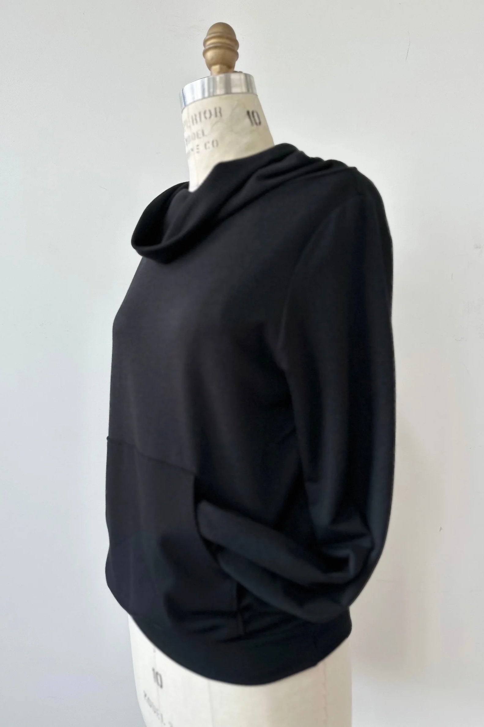 The Jullian Sweater by Kollontai in Black, with a cowl neck, long sleeves, and a kangaroo pocket, is shown on a mannequin in front of a white background. 
