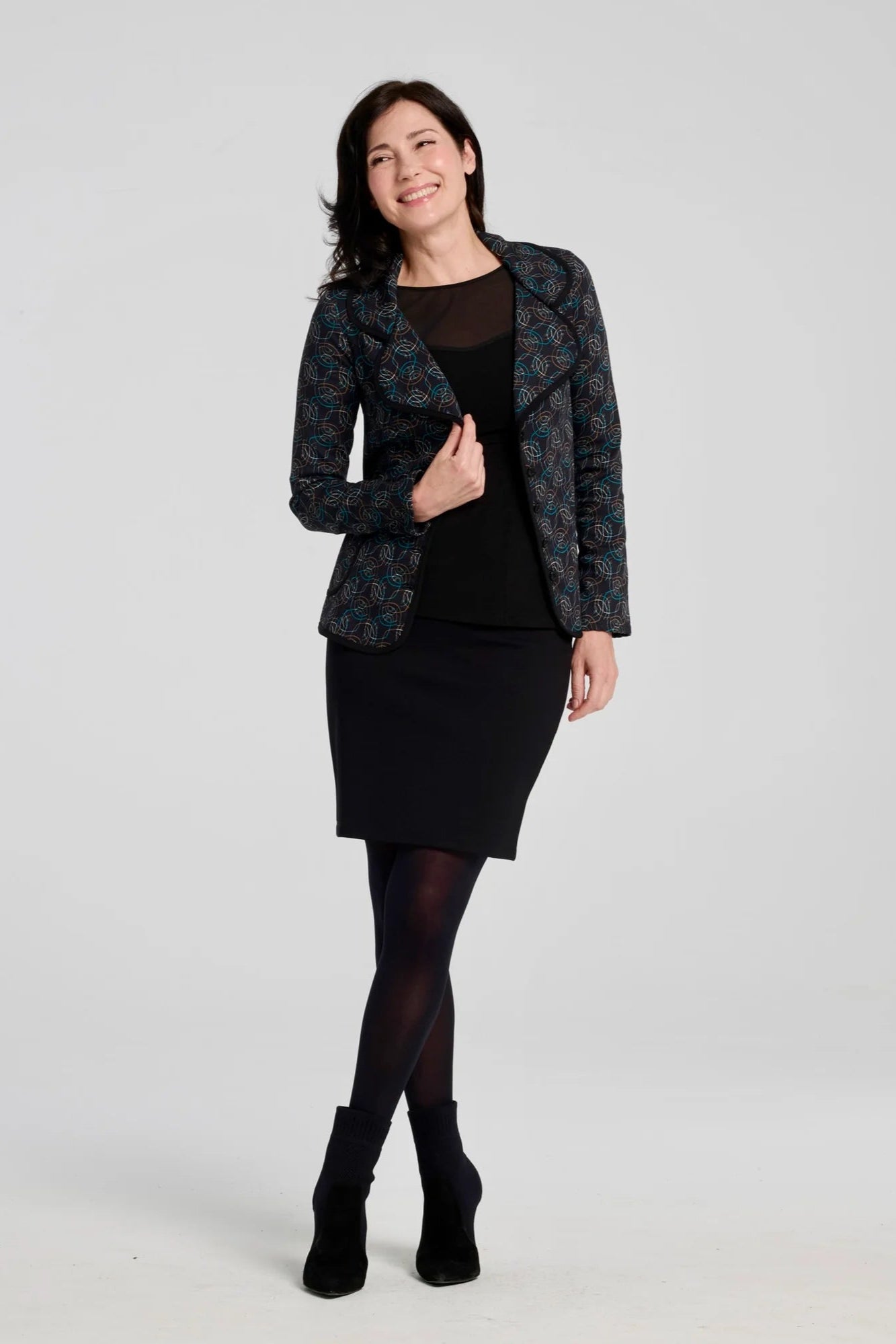 A woman wearing the Veneto Skirt by Kollontai in Black, a pencil skirt with a wide-pull on waistband, back slit, and hem that hits above the knee. She is wearing it with a black top and patterned and standing in front of a grey background. 