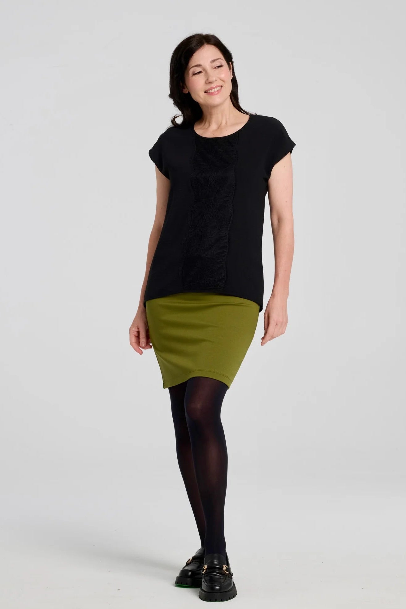 A woman wearing the Veneto Skirt by Kollontai in Granny Smith, a pencil skirt with a wide-pull on waistband, back slit, and hem that hits above the knee. She is wearing it with a black top and standing in front of a grey background. 