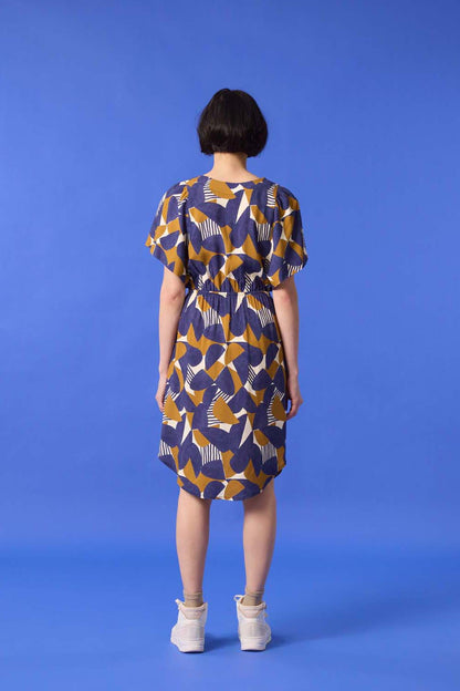 Back view of a  woman wearing the Gemma Dress by Cokluch in a Cumin half-moon print, standing in front of a blue background 