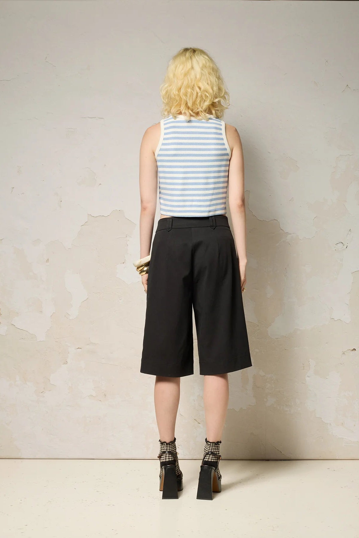 A woman wearing the Loke Shorts from Cokluch in Black, long Bermuda shorts with wide legs, a slightly dropped waist, and pointed belt loops. She is wearing them with a cropped striped tank top and standing in front of a grey wall. 