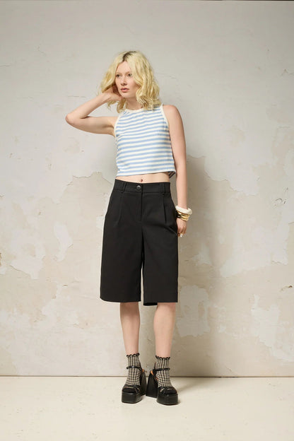 A woman wearing the Loke Shorts from Cokluch in Black, long Bermuda shorts with wide legs, a slightly dropped waist, and pointed belt loops. She is wearing them with a cropped striped tank top and standing in front of a grey wall. 