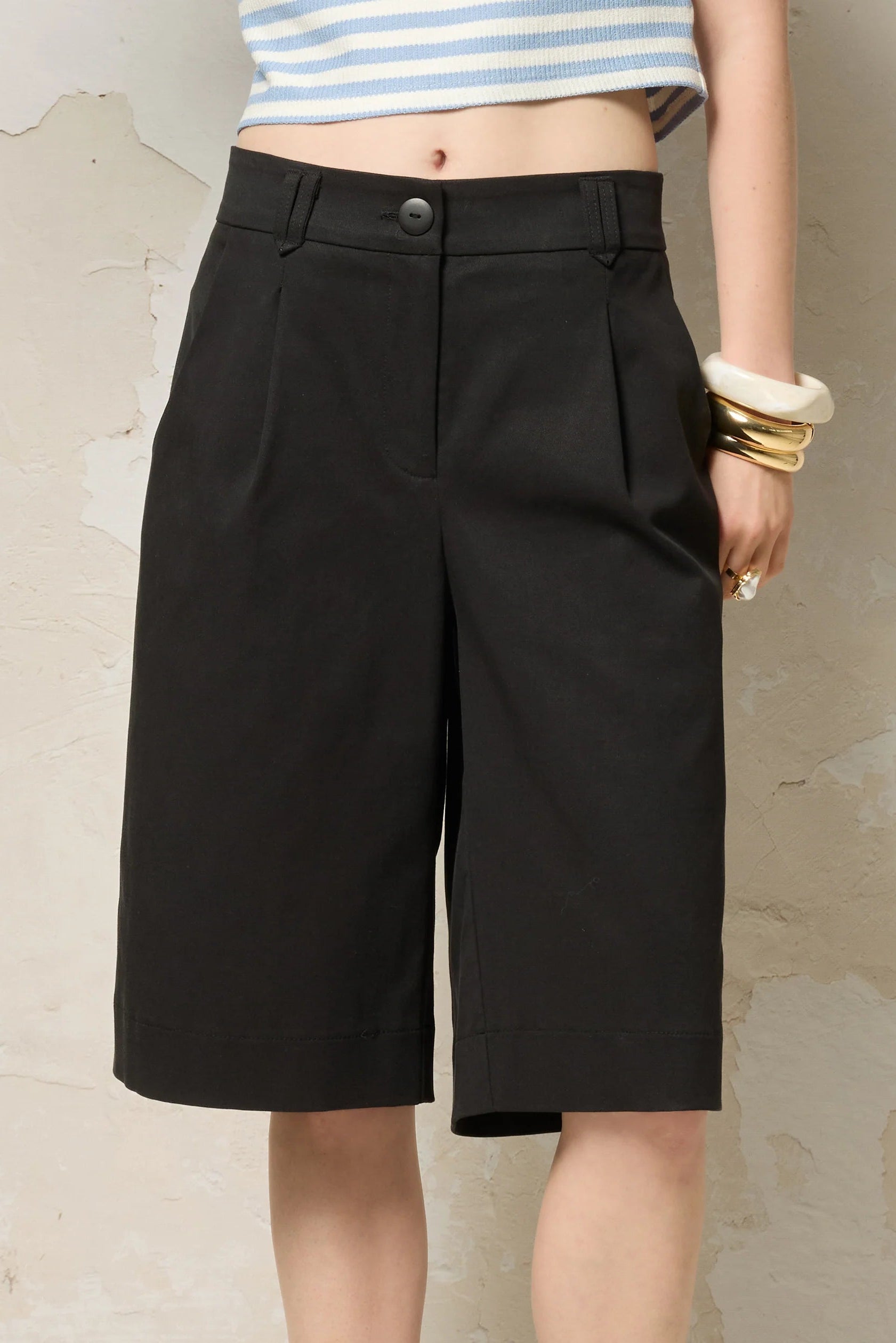 Waist down view of a woman wearing the Loke Shorts from Cokluch in Black, long Bermuda shorts with wide legs, a slightly dropped waist, and pointed belt loops. She is wearing them with a cropped striped top and standing in front of a grey wall. 