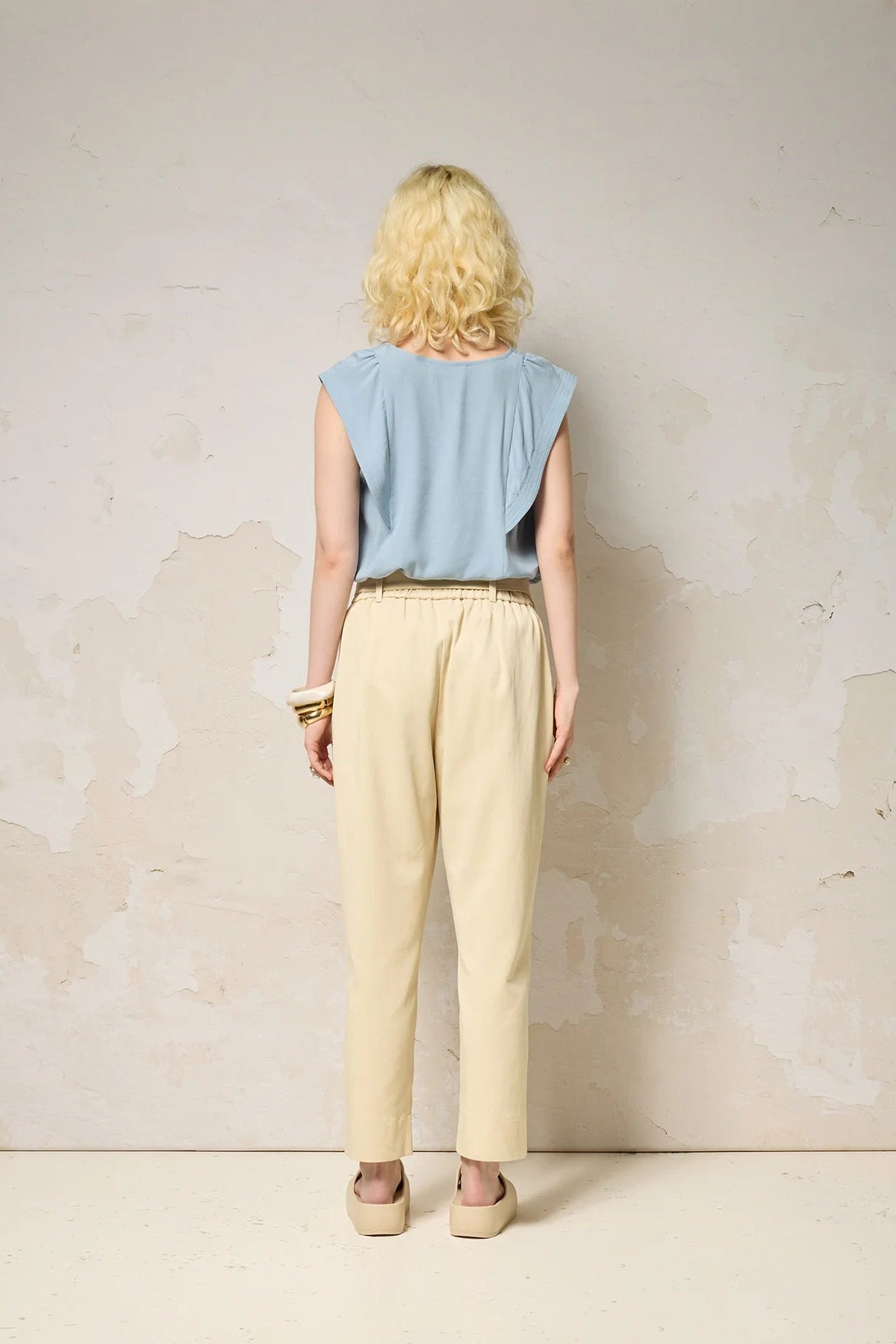 Back view of a woman wearing the Hima Top from Cokluch in Sky, a sleeveless top with shoulder ruffles, a round neck, and hip-length cut. She is wearing it with beige pants and standing against a textured background. 