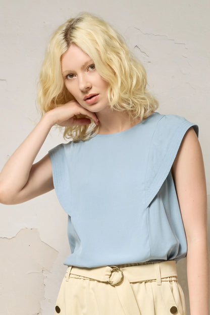 Waist up view of a woman wearing the Hima Top from Cokluch in Sky, a sleeveless top with shoulder ruffles, a round neck, and hip-length cut. She is wearing it with beige pants and standing against a textured background. 