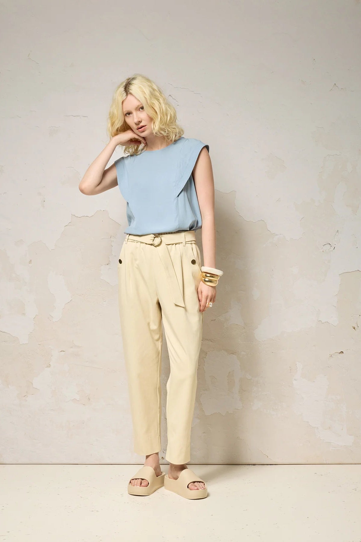  A woman wearing the Hima Top from Cokluch in Sky, a sleeveless top with shoulder ruffles, a round neck, and hip-length cut. She is wearing it with beige pants and standing against a textured background. 