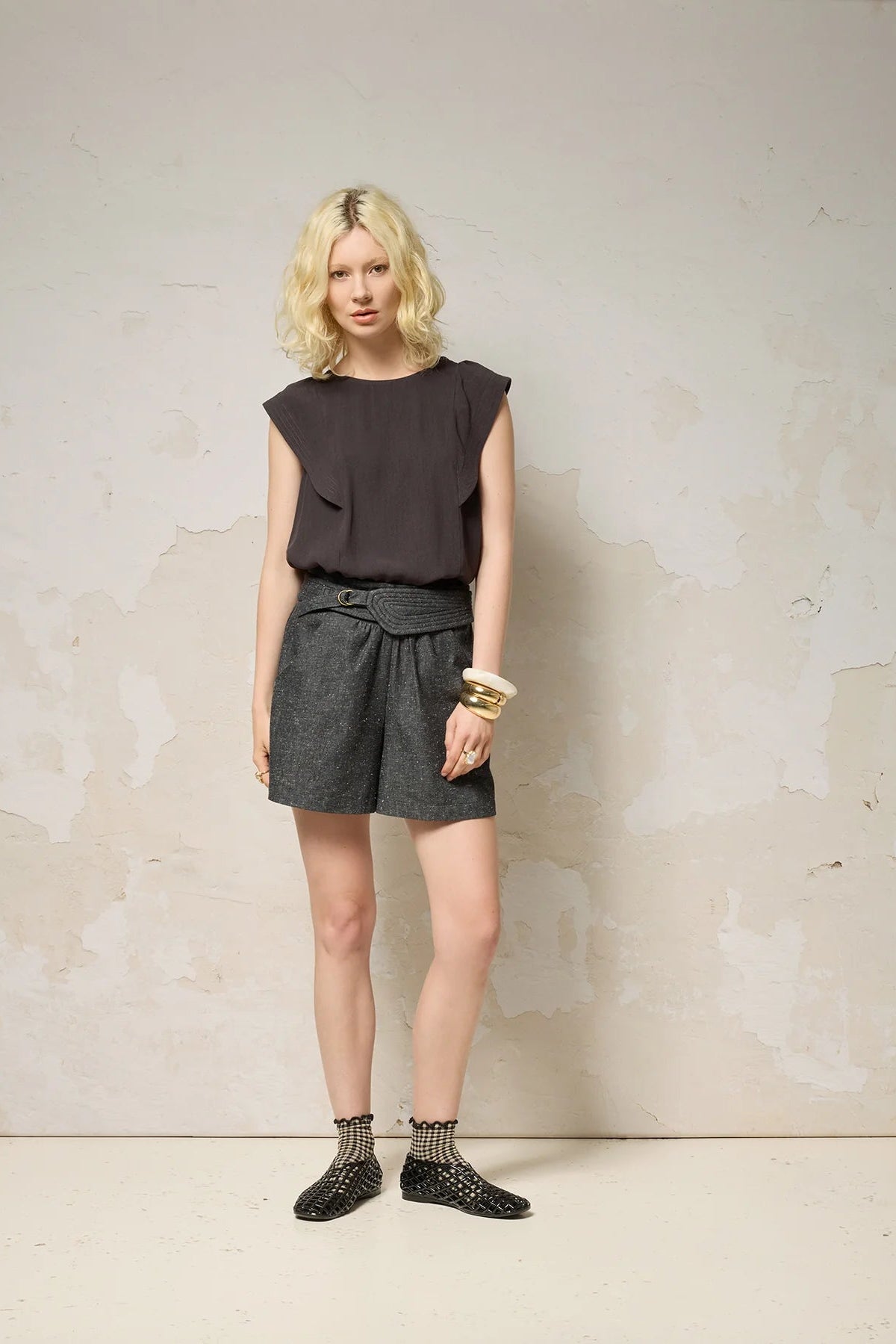  A woman wearing the Hima Top from Cokluch in Iron, a sleeveless top with shoulder ruffles, a round neck, and hip-length cut. She is wearing it with denim shorts and standing against a textured background. 