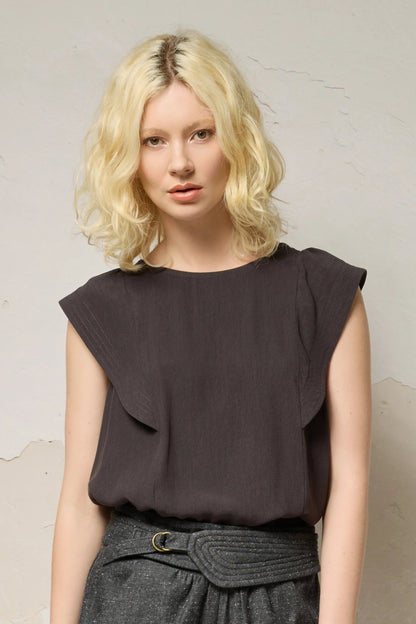 Waist up view of a woman wearing the Hima Top from Cokluch in Iron, a sleeveless top with shoulder ruffles, a round neck, and hip-length cut. She is wearing it with denim shorts and standing against a textured background. 