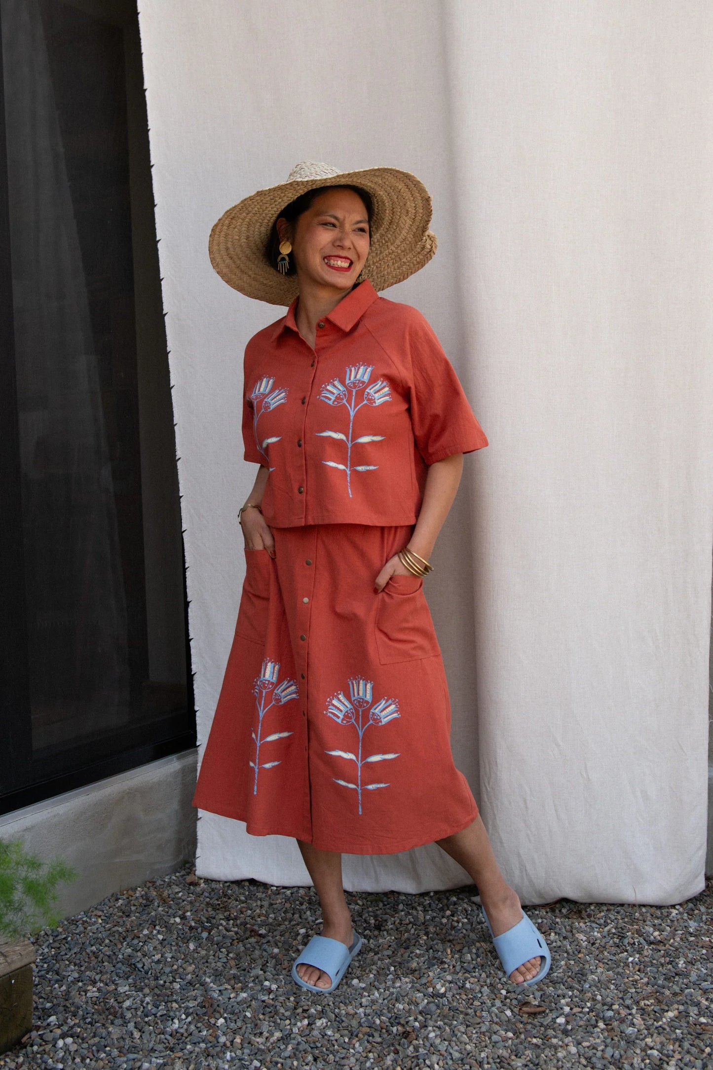 A woman wearing the Hosta Screen-printed Blouse by Kazak in Red with Pysanka, with hand-screen-printed flowers, mid-length raglan sleeves, a classic collar, and a button front. She is wearing it with a matching skirt and straw hat and standing outside by a white curtain. 