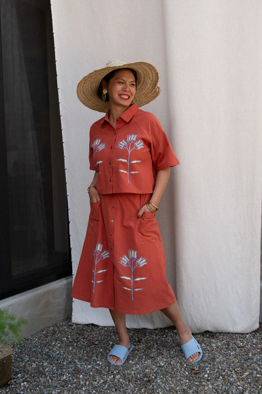 A woman wearing the Hosta Screen-printed Blouse by Kazak in Red with Pysanka, with hand-screen-printed flowers, mid-length raglan sleeves, a classic collar, and a button front. She is wearing it with a matching skirt and straw hat and standing outside by a white curtain. 