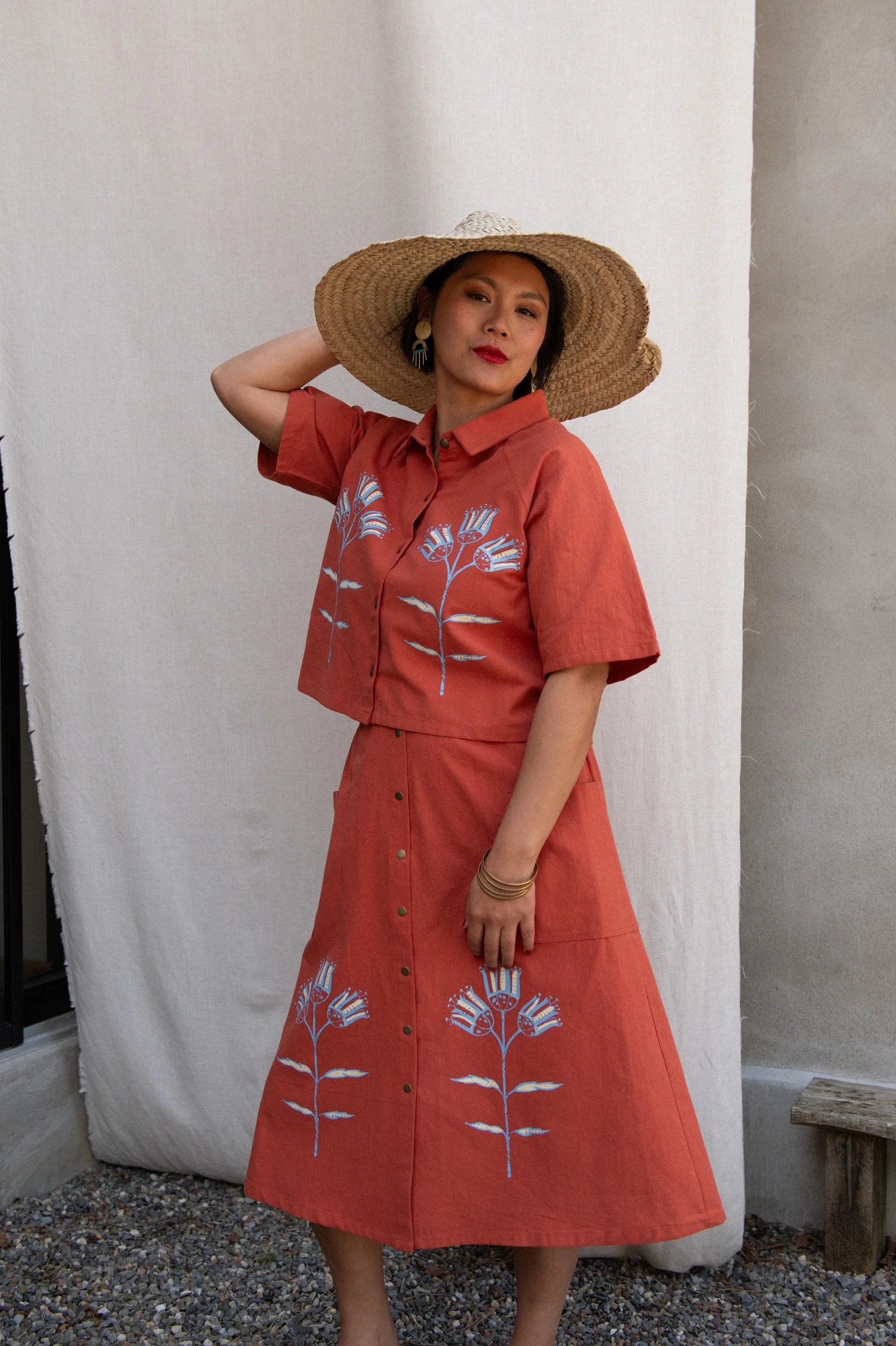 A woman wearing the Nymphea Screenprinted Skirt by Kazak in Red Pysanka with the matching Hosta top, she is standing outside in front of white curtain. 