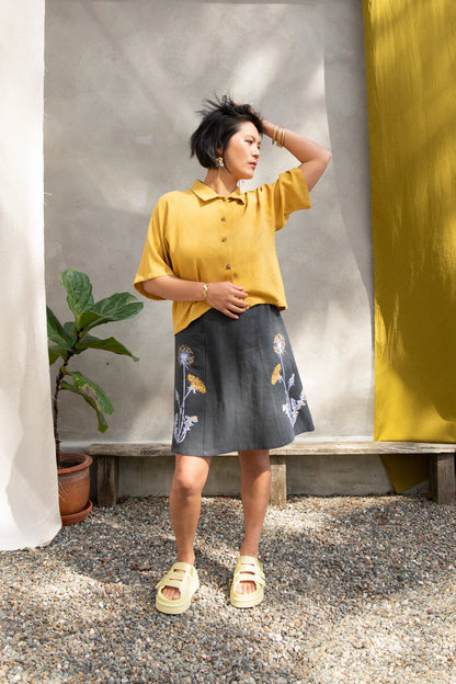 A woman wearing the Vicenze Screen-printed Skirt by Kazak in Black with Dandelions, an above the knee A-line skirt with hand-screened flowers, an elasticated waist, and large patch pockets. She is wearing it with the  Hosta shirt in Mustard and standing outside by a grey wall and white and yellow curtains.