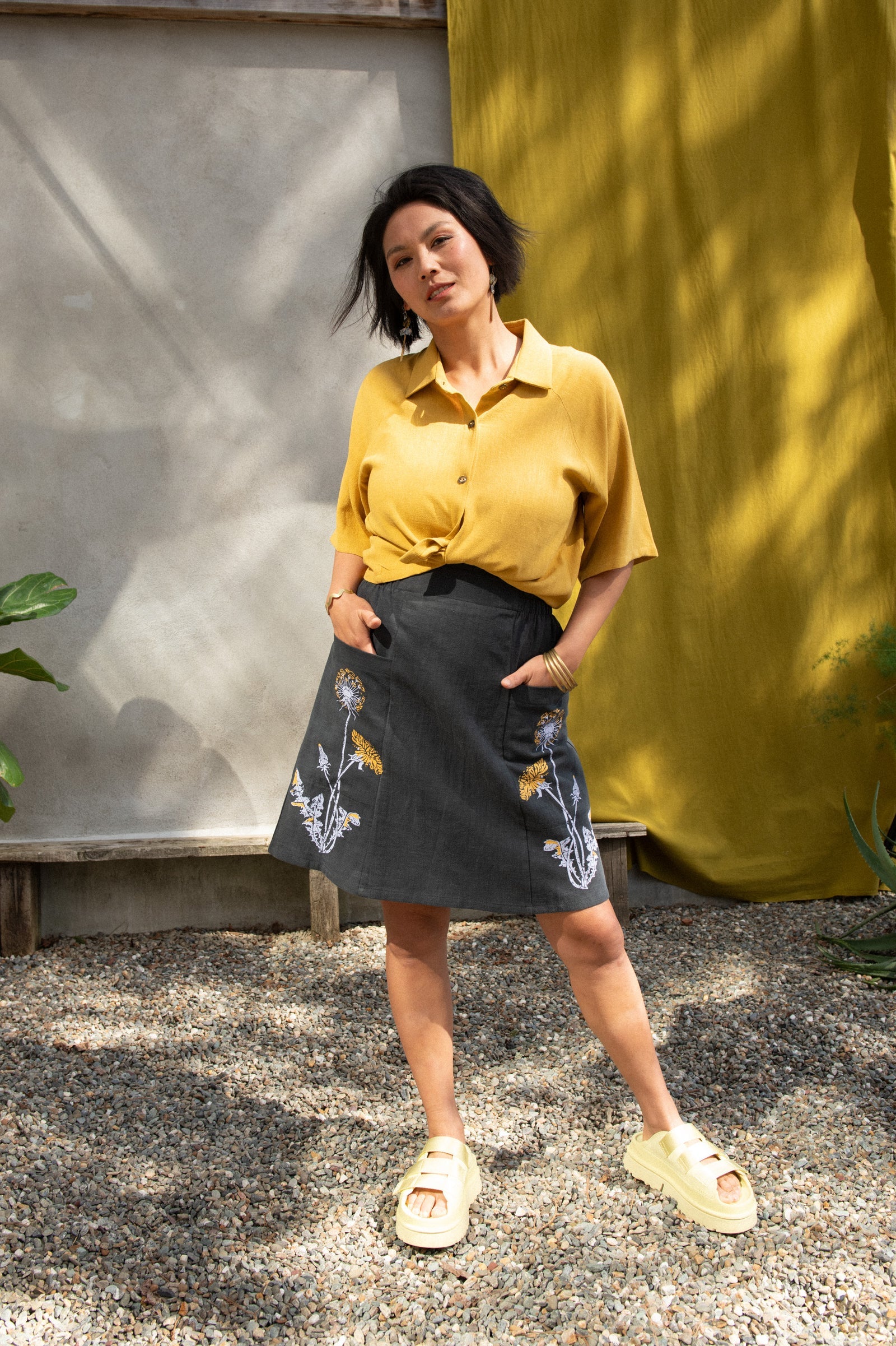 A woman wearing the Vicenze Screen-printed Skirt by Kazak in Black with Dandelions, an above the knee A-line skirt with hand-screened flowers, an elasticated waist, and large patch pockets. She is wearing it with the  Hosta shirt in Mustard and standing outside by a grey wall and white and yellow curtains.