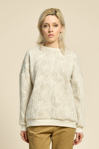 A woman wearing the Hotel Sweater by Cokluch in Natural, a loose fitting round necked sweater in Crinkle Max fabric. She is wearing it with yellow pants and standing in front of a beige background. 