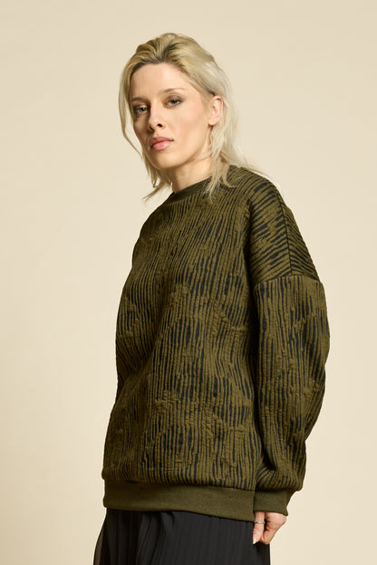 A woman wearing the Hotel Sweater by Cokluch in Olive, a loose fitting round necked sweater in Crinkle Max fabric. She is wearing it with black pants and standing in front of a beige background. 