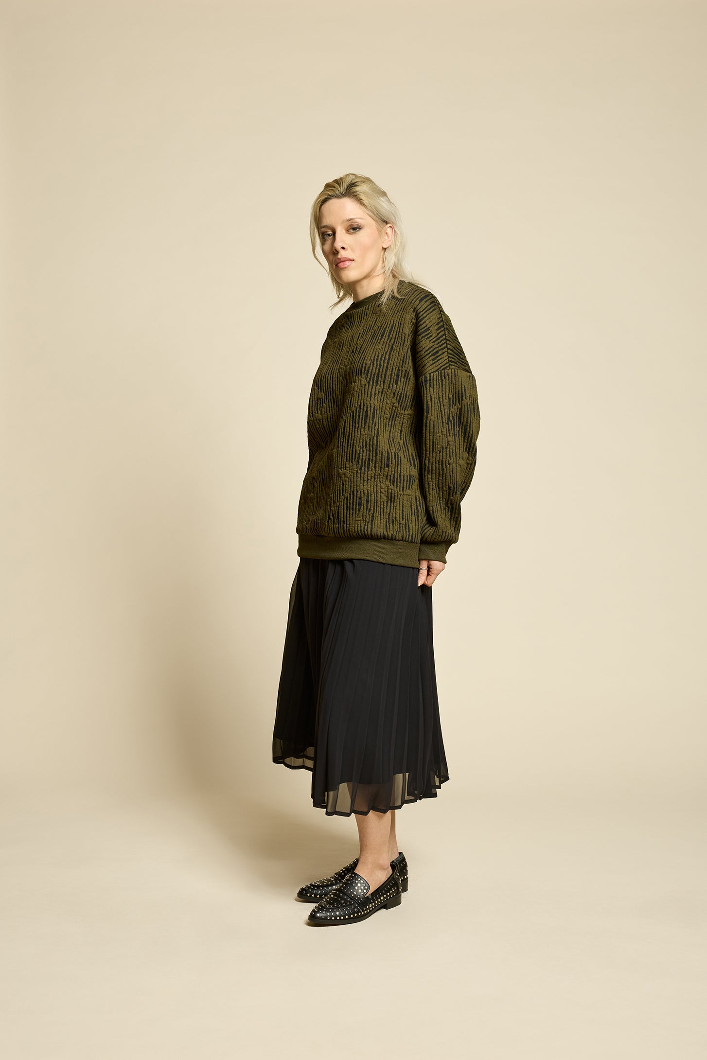 A woman wearing the Hotel Sweater by Cokluch in Olive, a loose fitting round necked sweater in Crinkle Max fabric. She is wearing it with a bladck skirt and standing in front of a beige background. 