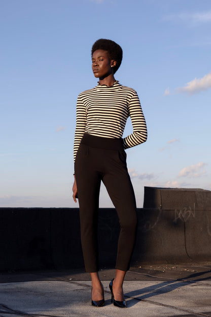 A woman wearing the Ponte di Roma Larimar Pants by Eve Lavoie in Black, slim-fitting pull-on pants with a wide waistband, front pleats, and an ankle-length hem with a side slit detail. She is standing outside and wearing a striped turtleneck. 