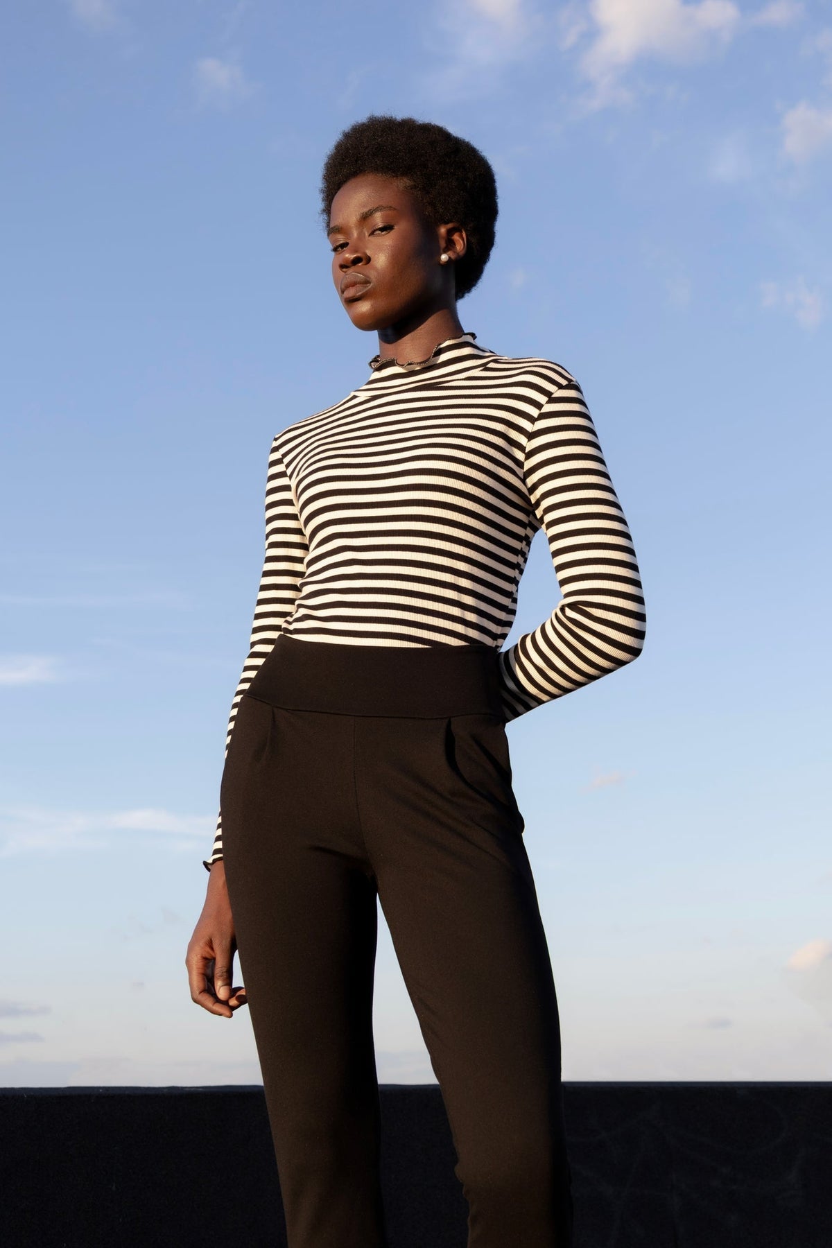 A woman wearing the Ponte di Roma Larimar Pants by Eve Lavoie in Black, slim-fitting pull-on pants with a wide waistband, front pleats, and an ankle-length hem with a side slit detail. She is standing outside and wearing a striped turtleneck. 