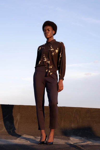 A woman wearing the Ponte di Roma Larimar Pants by Eve Lavoie in Navy, slim-fitting pull-on pants with a wide waistband, front pleats, and an ankle-length hem with a side slit detail. She is standing outside and wearing a floral blouse. 