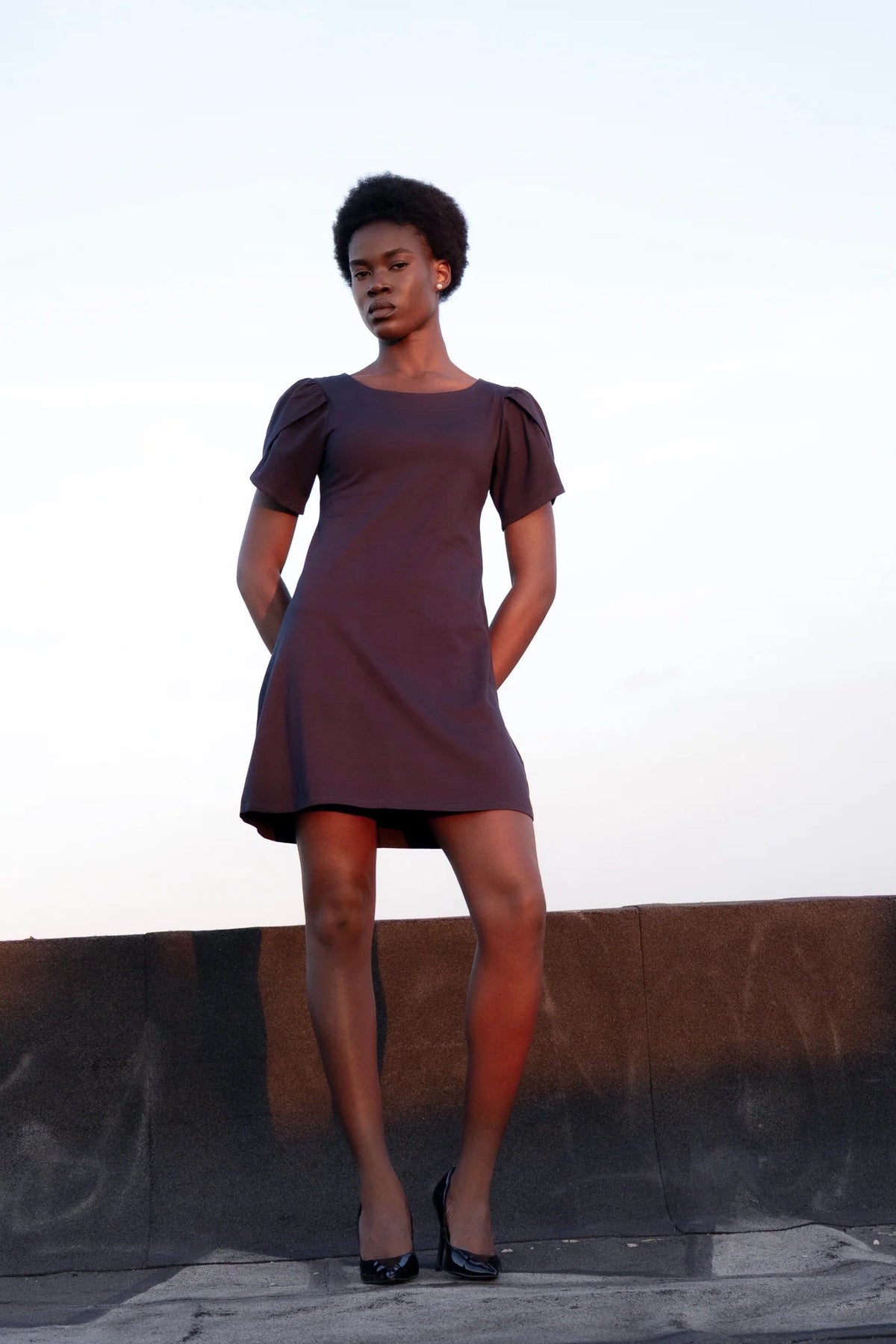 A woman wearing the Opal Dress by Eve Lavoie in Navy, a short dress with tulip sleeves, and an A-line shape. She's standing outdoors by a low concrete wall. 
