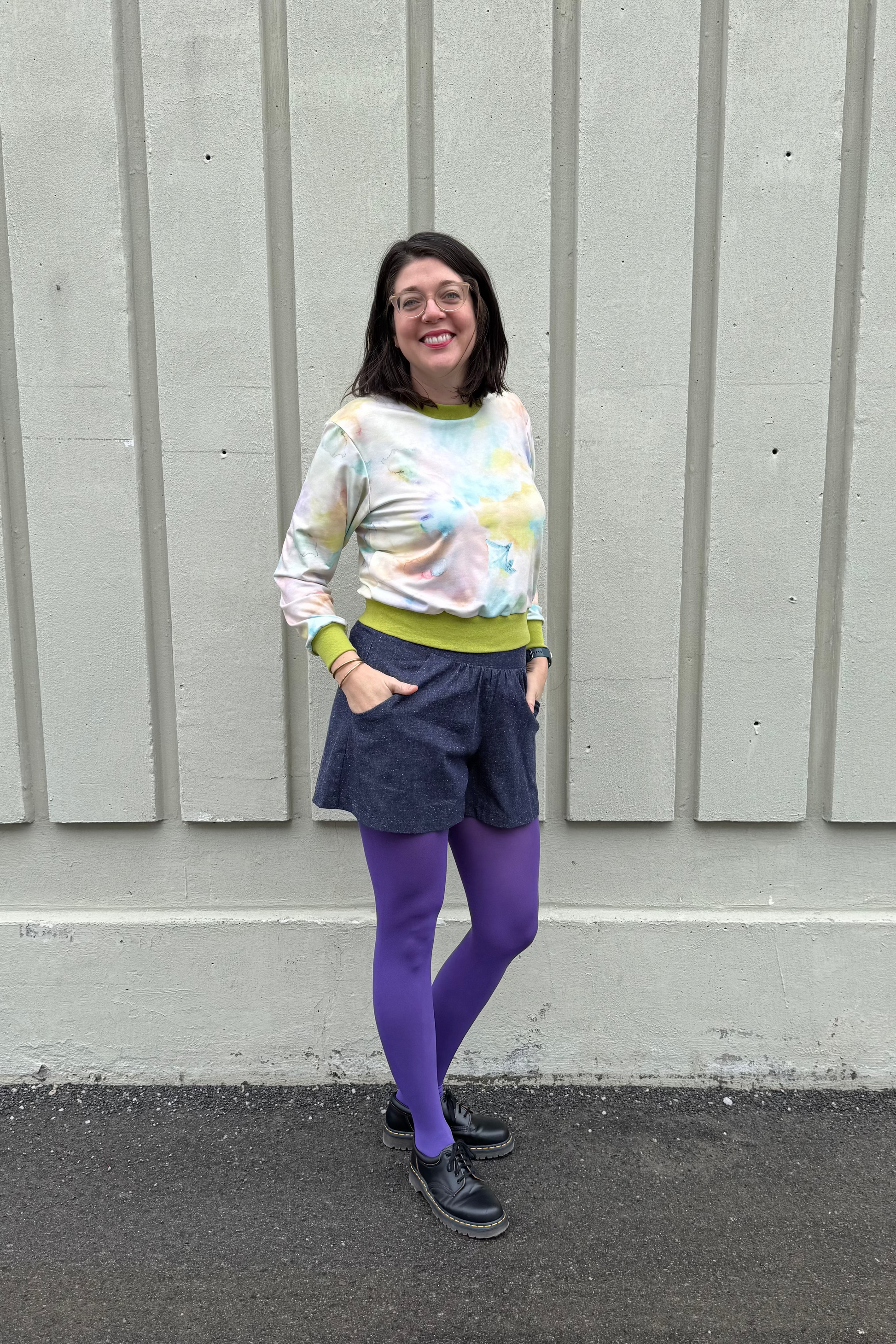 A woman wearing the Wanda E26 Sweater by Slak in Watercolour print, with a crew neck, long sleeves, and contrasting green ribbing at the cuffs, collar, and hem. She is wearing it with navy shorts and purple tights and standing outside in front of panelled wall. 