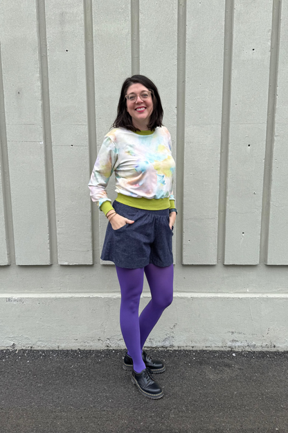 A woman wearing the Wanda E26 Sweater by Slak in Watercolour print, with a crew neck, long sleeves, and contrasting green ribbing at the cuffs, collar, and hem. She is wearing it with navy shorts and purple tights and standing outside in front of panelled wall. 