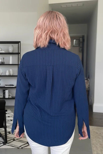 Back view of a woman wearing the Blue Striped Blouse by Dotty, a classic button front shirt with a collar and extra wide cuffs. She is wering it with white pants and standing in a room with shelves.