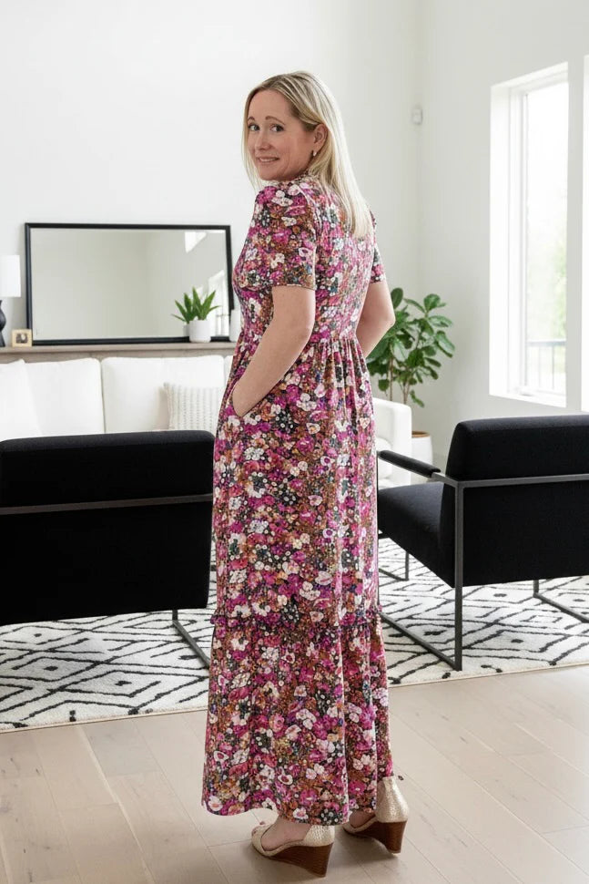 Back view of a woman wearing the Petal Print by Dotty in Pink/Blue floral,  a maxi dress with short flutter sleeves, a split V-neck and ruffled collar, with a gathered waist and A-line skirt. She is standing in a modern living room.
