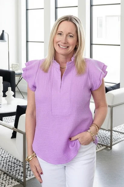 A woman wearing the Purple Cotton Blouse by Dotty, with short double ruffled sleeves, a split v-neck, ruffle-banded colour and rounded hi-low hem. She is wearing it with white pants in an indoor setting. 