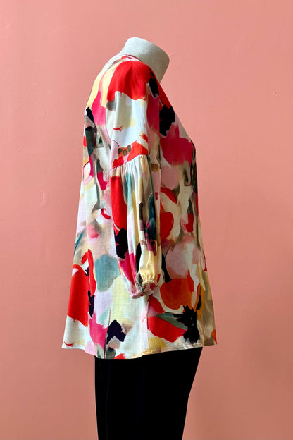 A side view of the Grace Blouse by Pure Essence in Coral Meadow, with a v-placket neck with buttons and 7/8 gathered sleeves, is shown with black pants on a mannequin in front of a coral wall. 