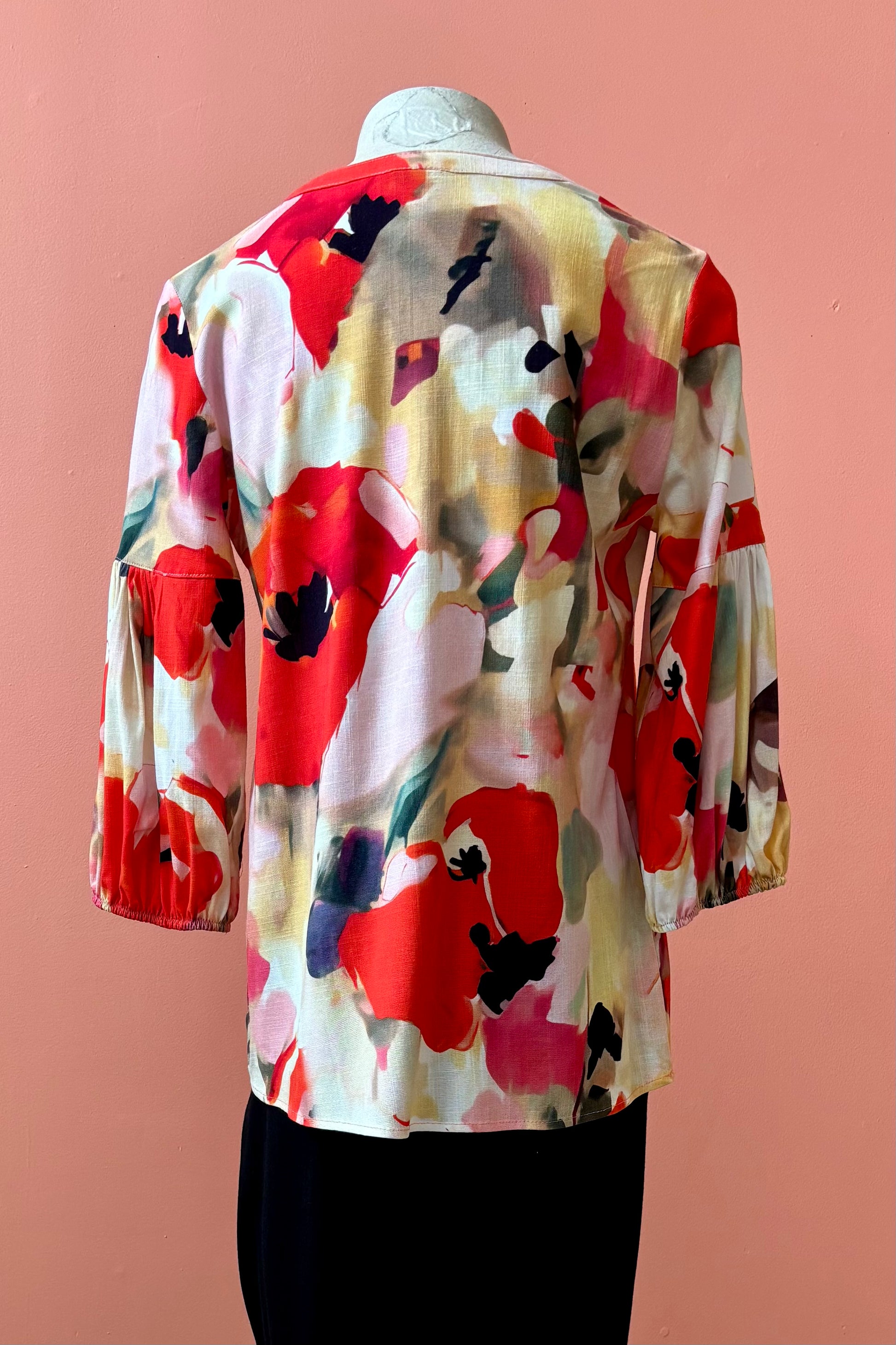 A back view of the Grace Blouse by Pure Essence in Coral Meadow, with a v-placket neck with buttons and 7/8 gathered sleeves, is shown with black pants on a mannequin in front of a coral wall. 