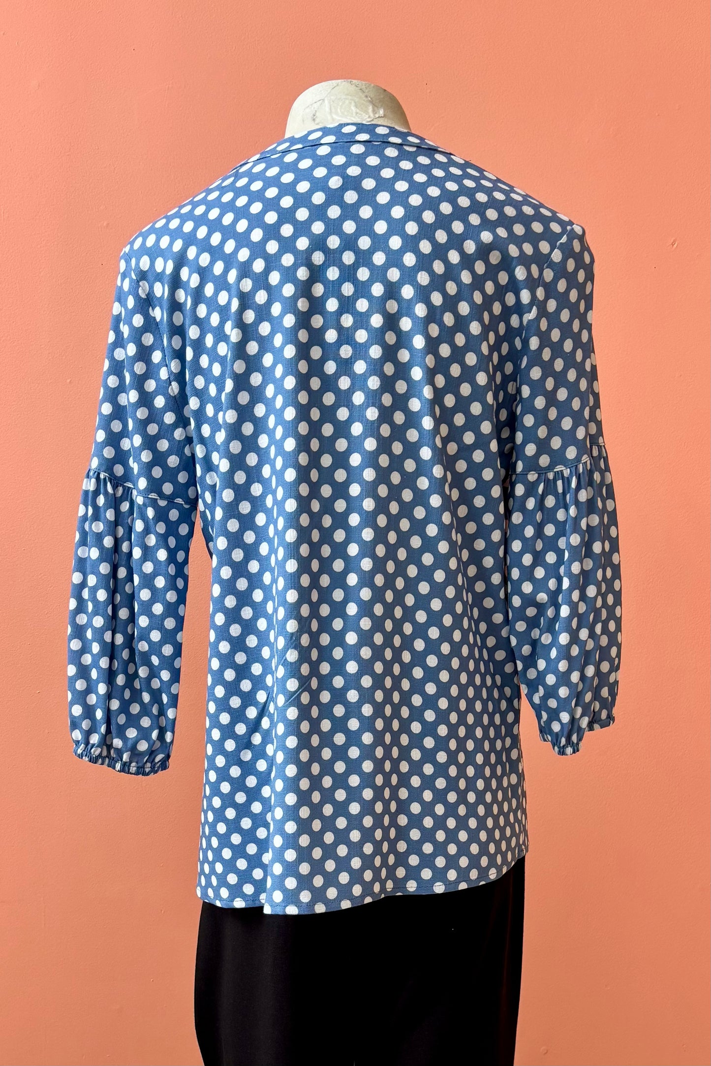 A back view of the Grace Blouse by Pure Essence in Chambray Dot, with a v-placket neck with buttons and 7/8 gathered sleeves, is shown with black pants on a mannequin in front of a coral wall. 