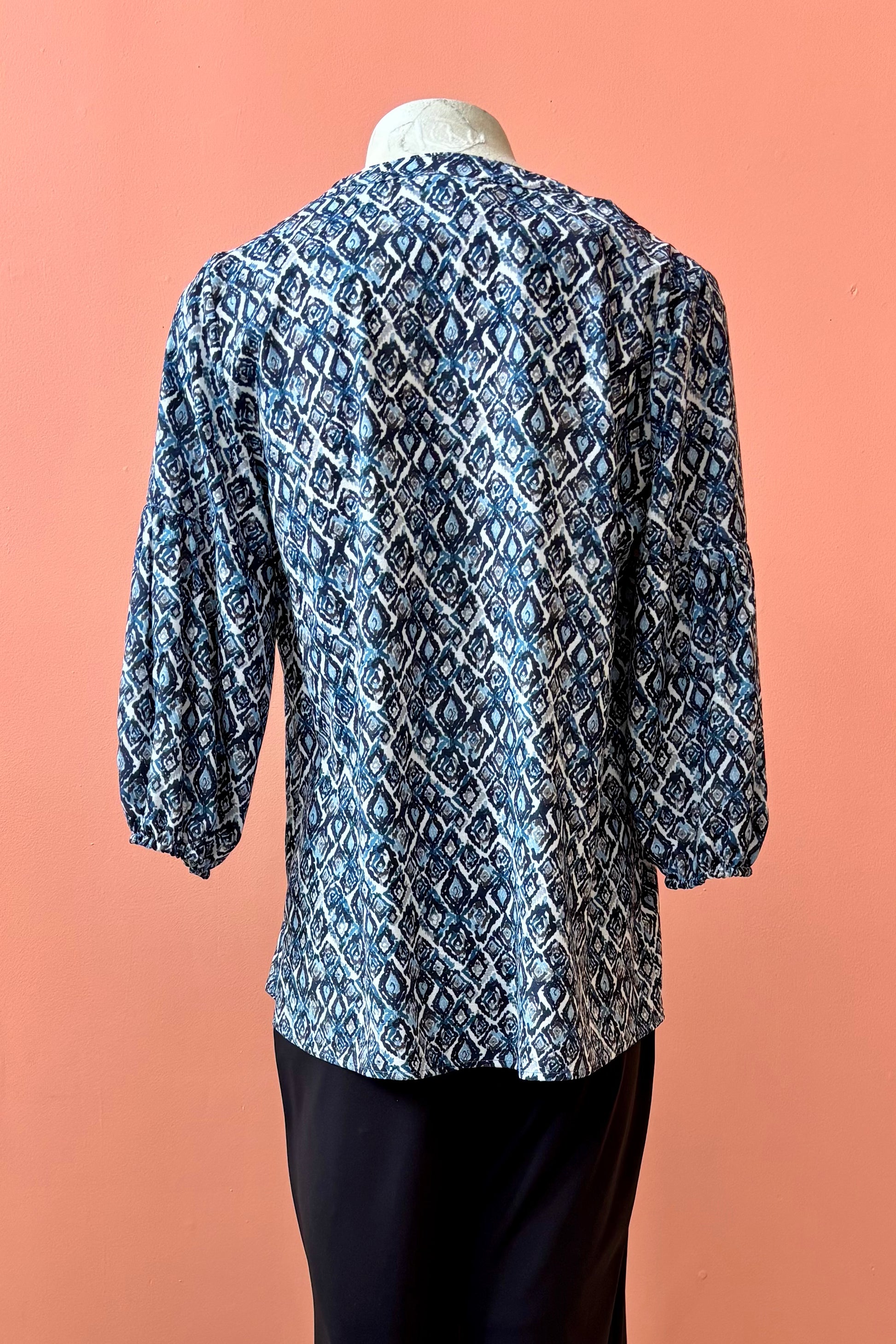 A back view of the Grace Blouse by Pure Essence in Blue Lattice, with a v-placket neck with buttons and 7/8 gathered sleeves, is shown with black pants on a mannequin in front of a coral wall. 
