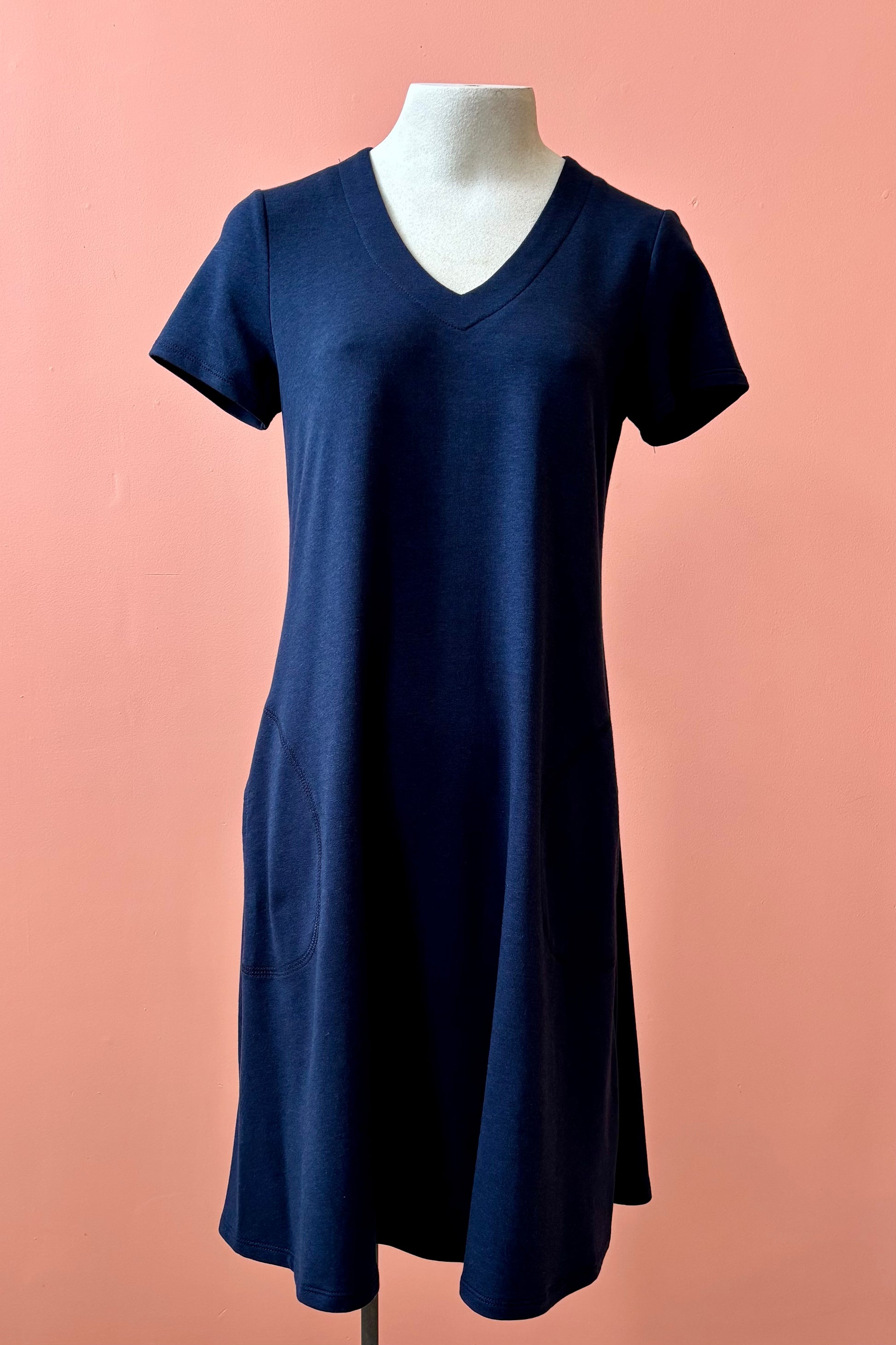 The Gaia Dress by Pure Essence in Navy, a short sleeved knee length dress with a V-neck, A-line shape, and pockets, is shown on a mannequin against a coral wall.