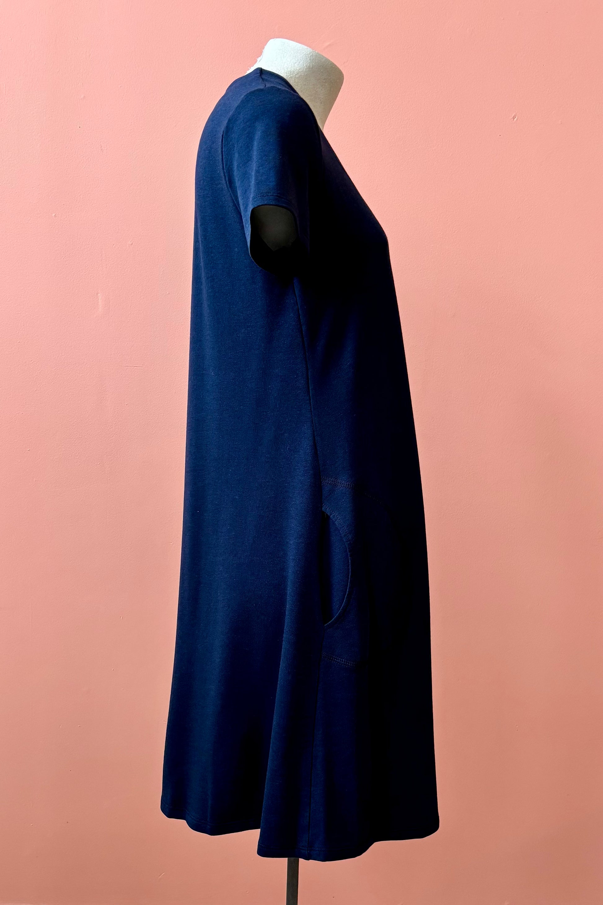 A side view of the Gaia Dress by Pure Essence in Navy, a short sleeved knee length dress with a V-neck, A-line shape, and pockets, is shown on a mannequin against a coral wall.