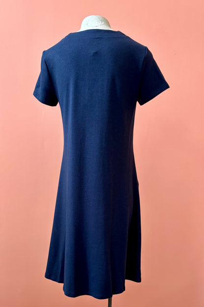 A back view of the Gaia Dress by Pure Essence in Navy, a short sleeved knee length dress with a V-neck, A-line shape, and pockets, is shown on a mannequin against a coral wall.